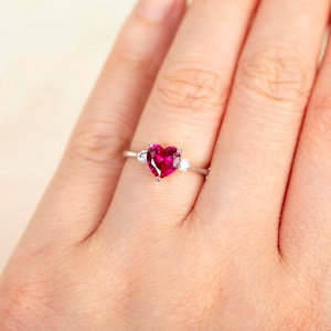 K&ouml;nnte beinhalten: Ein silberner Ring mit einem gro&szlig;en herzf&ouml;rmigen Rubin-Edelstein und zwei kleineren runden wei&szlig;en Edelsteinen auf der Ringschiene.