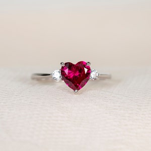 K&ouml;nnte beinhalten: Ein silberner Ring mit einem gro&szlig;en herzf&ouml;rmigen Rubin-Edelstein und zwei kleineren runden, klaren Edelsteinen auf der Ringschiene.