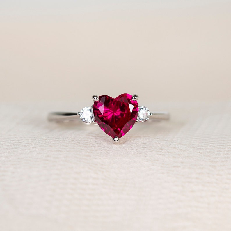 K&ouml;nnte beinhalten: Ein silberner Ring mit einem gro&szlig;en herzf&ouml;rmigen Rubin-Edelstein und zwei kleineren runden, klaren Edelsteinen auf der Ringschiene.