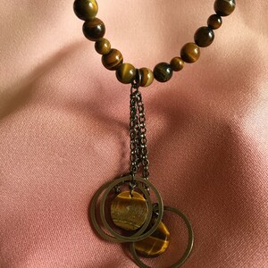 May include: A necklace with tiger's eye beads and two circular pendants. Each pendant has a tiger's eye stone. The necklace hangs on a pink fabric. The chain and pendants are made of a dark metal.