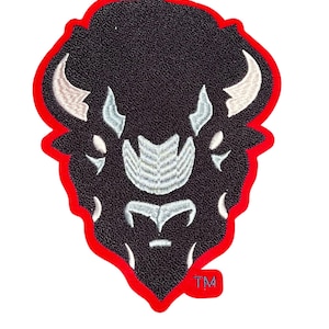 May include: Embroidered patch featuring a buffalo head design. The patch has a black textured background with white and light blue details. The buffalo head is outlined in red, with the letters "TM" in the lower right corner.