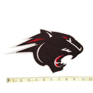 May include: A black panther head patch with red accents on the eyes and mouth. The patch has a textured surface and a white outline. A measuring tape is placed below the patch, showing a length of approximately 25 cm.