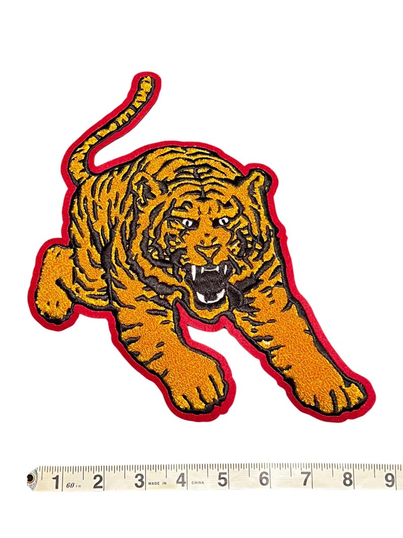 May include: Embroidered tiger patch in shades of orange and black, outlined with a red border. The tiger is depicted in a dynamic pose with its mouth open, showcasing its teeth. The patch is designed for application on clothing or accessories.