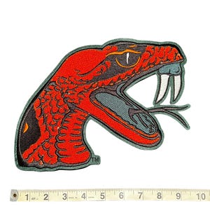 May include: A red and black embroidered snake patch with an open mouth, fangs, and a forked tongue. The patch has a green border and is approximately 4 inches wide.