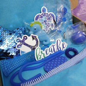 May include: A selection of blue sensory toys and stickers in a box. Items include a sequin pillow, a seahorse, a turtle sticker, a whale, a finger massager, and a blue brush. The word "breathe" is written on a sticker.