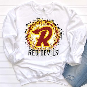 May include: White sweatshirt with a red and yellow stylized letter "R" with a red arrow inside the letter. The letter is surrounded by a yellow, red, and black splatter pattern. The text "RED DEVILS" is below the letter.
