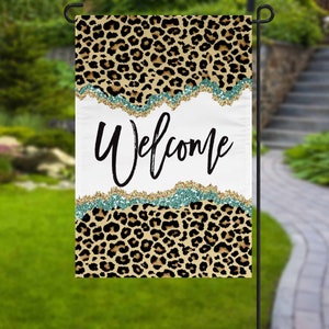 May include: A garden flag with a leopard print design and the word "Welcome" in black script. The flag has a white center with teal and gold glitter accents. The top and bottom of the flag feature a brown and black leopard print.