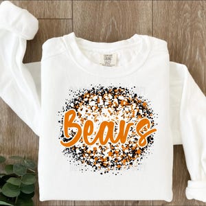 May include: White long-sleeve shirt with the word "Bears" in orange script, surrounded by a speckled design of black, white, and orange dots. The shirt is folded on a wooden surface.