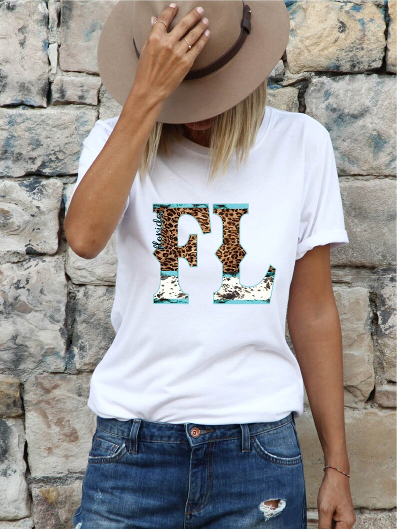 May include: White t-shirt with a graphic design featuring the letters "FL" in a leopard print pattern, with turquoise accents. The word "Florida" is printed vertically on the left side of the "F". The model is wearing blue denim shorts.
