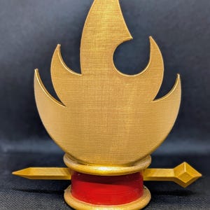 May include: A gold-colored decorative object with a flame-like design. The object has a red base and a horizontal gold bar with a diamond-shaped end. The flame design has a textured surface.