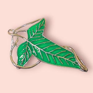 May include: A green leaf-shaped brooch with gold-colored trim and a silver-colored vine design. The brooch has a glossy finish and is set against a light pink background. The leaf has detailed veining.