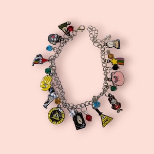 May include: A silver charm bracelet with multiple charms featuring characters and symbols from the TV show Gravity Falls. The charms include a pig, a hand, a triangle, a star, a rainbow, a gnome, a character with a hat, and a character with a baseball cap.