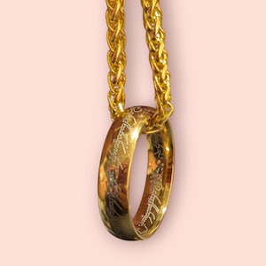 May include: A gold ring with engraved text, hanging from a gold chain. The ring is circular and polished, and the chain has a woven texture. The background is a soft pink. The ring is likely a piece of jewellery.