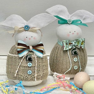 May include: Two handmade bunny sock dolls with brown knitted sweaters and white fabric ears. The bunny on the left has a blue and brown bow and a green Spartan helmet charm. The bunny on the right has a floral print bow and a green Spartan helmet charm.