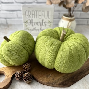 May include: Two green knitted pumpkins on a wooden cutting board. The larger pumpkin has a wooden stem. The smaller pumpkin is to the left of the larger pumpkin. The pumpkins are on a wooden cutting board with a decorative handle. The background is a white brick wall with a sign that says "Grateful Hearts Gather Here".