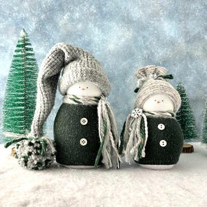 May include: Two handmade snowman figures with gray knit hats and scarves, wearing dark green sweater bodies with button details. The snowmen are decorated with a pom-pom and snowflake accents, set against a wintery backdrop with miniature Christmas trees.