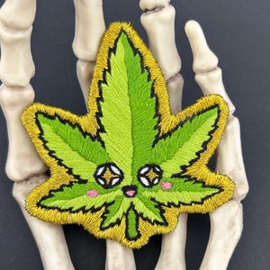 Op de afbeelding: Een geborduurde patch in de vorm van een cannabisblad met een felgroen midden, een dikke goudgele rand en een zwarte omlijning. Het blad heeft een cartoongezicht met twee stervormige ogen, een kleine roze mond en twee roze wangen.