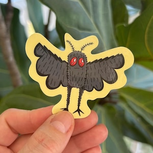 May include: A yellow sticker featuring a cartoon moth with black wings, a fuzzy body, and red eyes. The moth has small antennae and thin black legs. The sticker is held against a background of green leaves.