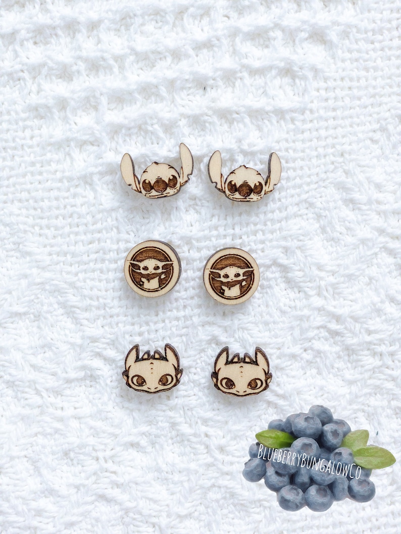 May include: A set of six wooden earrings featuring cartoon characters. The earrings are in the shape of Stitch, Baby Yoda, and Toothless from the movie How to Train Your Dragon. The earrings are on a white background.