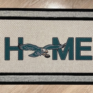 Könnte beinhalten: Eine Fußmatte mit schwarzem Rahmen und grauem Rand. Die Fußmatte hat einen weißen Hintergrund mit dem Wort "HOME" in grünen Buchstaben. Ein grüner Adler mit einem Fußball fliegt über dem Wort "HOME".