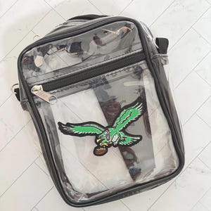 May include: Clear crossbody bag with black trim and zipper closure. Features a green and white embroidered eagle patch holding a football. Includes a black adjustable shoulder strap.