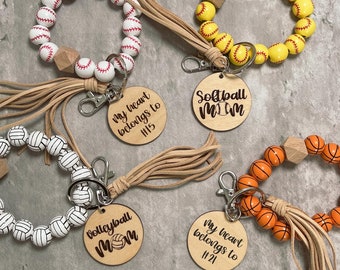 May include: A collection of beaded wrist keychains. The keychains feature beads designed as baseballs, softballs, volleyballs, and basketballs. Each keychain has a wooden tag with text such as "Softball Mom" and "My heart belongs to #15".