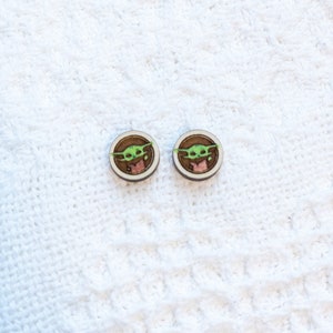 May include: A pair of wooden earrings with a green character from the Star Wars franchise, The Mandalorian, known as "The Child" or "Baby Yoda".