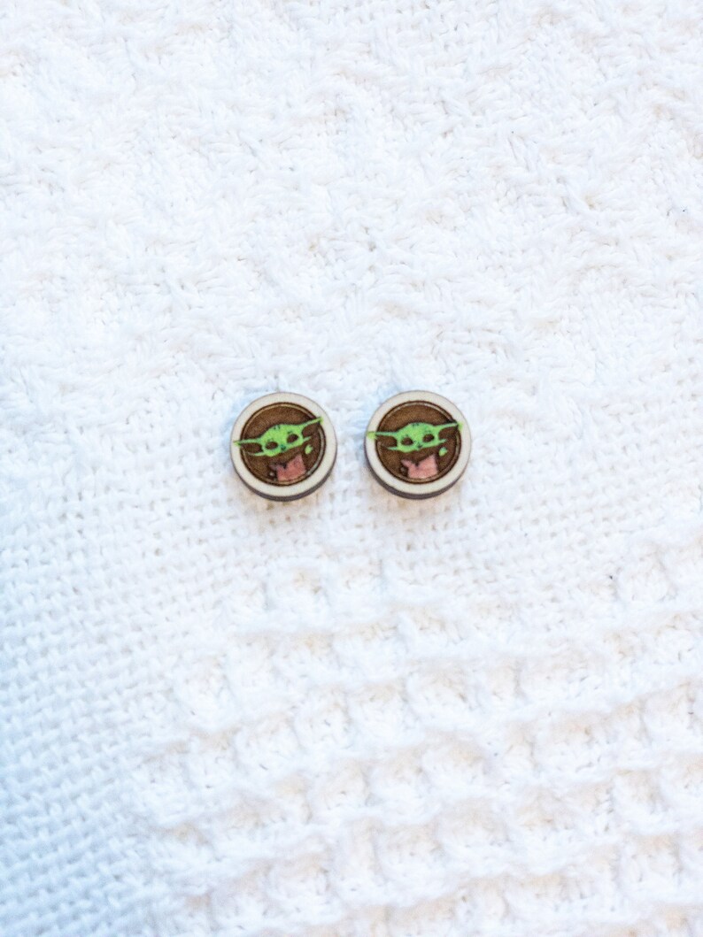 May include: A pair of wooden earrings with a green character from the Star Wars franchise, The Mandalorian, known as "The Child" or "Baby Yoda".