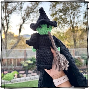 Könnte beinhalten: Eine gehäkelte Hexenpuppe mit grünem Gesicht, schwarzem Kleid und braunem Besen. Die Puppe wird von einer Hand mit einem rosafarbenen Armband gehalten.