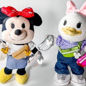 May include: Two plush Disney character dolls, Minnie Mouse and Daisy Duck, each holding a small purse. Minnie Mouse wears a red bow, denim skirt, and yellow shoes. Daisy Duck wears a pink bow, jeans, and white sneakers. Both dolls have purses.