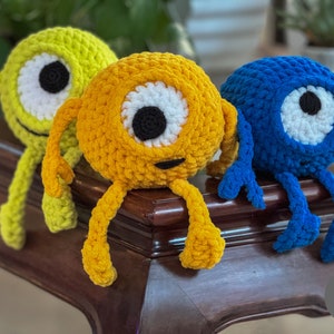 May include: Three crocheted plush toys with one large eye each. The toys are yellow, blue, and green and are sitting on a wooden table.