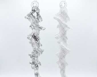 May include: Two hanging icicle ornaments made of clear and white material. The left icicle is transparent with embedded silver glitter accents, while the right icicle is opaque white with a dense coating of fine silver glitter throughout its surface.