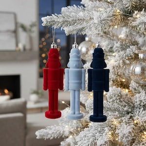 May include: Three nutcracker ornaments hanging from a flocked Christmas tree. The ornaments are made of a soft, velvet-like material in solid red, light blue, and dark navy blue colors, each featuring a silver cap and a thin hanging string.
