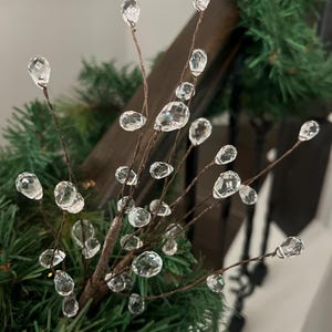 May include: A decorative floral pick featuring thin, brown wire stems adorned with multiple clear, faceted, teardrop-shaped acrylic beads. The stems branch out from a central point, creating a spray of crystal-like accents.