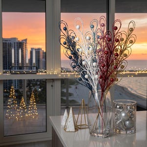 May include: A collection of decorative floral sprays featuring glittery wire loops and small spheres in blue, white, red, and gold. These sprays are arranged in a clear glass vase next to a glass container filled with silver ornaments.
