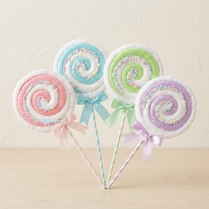 May include: Four round lollipop decorations featuring spiral patterns in pink, blue, green, and purple. Each lollipop is covered in white glitter and small colorful sprinkles, finished with a matching satin ribbon bow tied to a striped stick.