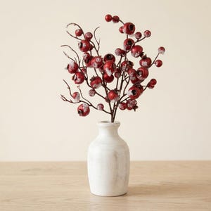 May include: A small white ceramic vase with a distressed finish holds a decorative branch featuring numerous round red berries. Each berry is accented with a dusting of white artificial snow, creating a textured appearance on the dark brown stems.