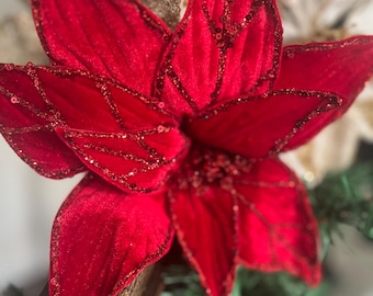 Könnte beinhalten: Eine leuchtend rote Weihnachtsstern-Blütenverzierung mit samtig strukturierten Blütenblättern. Die Blätter sind mit komplizierten, funkelnden roten Paillettenadern und einer zentralen Ansammlung roter Pailletten verziert. Vor einem unscharfen Hintergrund.