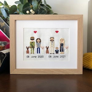 Può includere: Un ricamo a punto croce incorniciato con un ritratto di famiglia di quattro persone e due cani. Le persone sono raffigurate in uno stile di cartone animato stilizzato. Le date "08 giugno 2020" e "08 giugno 2021" sono scritte sotto le figure.