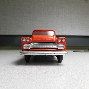 May include: A bright red toy pickup truck with chrome detailing on the front grill and headlights. The toy truck has black wheels and a classic vintage design, set against a neutral background.