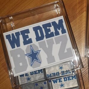 May include: A set of dominoes in a clear plastic case. The dominoes are gray with blue dots and have the words "We Dem Boyz" and a blue star on them.