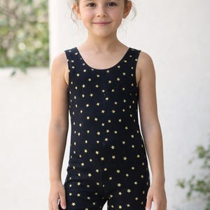 May include: A black romper with a scoop neck and short legs, covered in small gold stars. The child's outfit is made of a soft, stretchy fabric. The girl is smiling and looking at the camera.