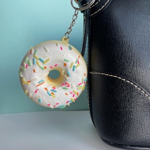 Op de afbeelding: Een witte donut sleutelhanger met kleurrijke sprinkels bevestigd aan een zwarte handtas.