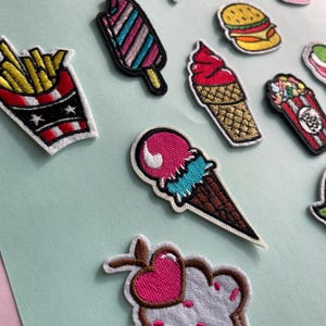 May include: Assortment of embroidered food patches, including a french fry container, ice cream cone, popsicle, pizza slice, cupcake, and hamburger. The patches feature vibrant colors and detailed stitching, perfect for adding flair to clothing or accessories.