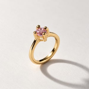 Könnte beinhalten: Ein goldener Ring mit einem einzelnen, rund geschliffenen rosa Edelstein in einer Vier-Krappen-Fassung. Die Ringschiene ist glatt und poliert und zeigt einen hellen Gelbgoldton, der mit dem zarten Rosaton des Steins kontrastiert.