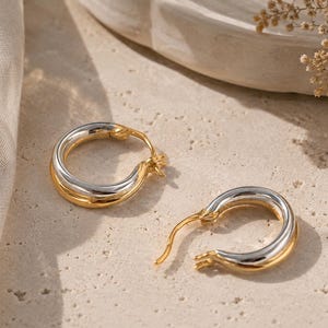 May include: A pair of small hoop earrings featuring a two-tone design with a polished silver-colored inner band and a smooth gold-colored outer band. The earrings have a hinged post closure mechanism and a rounded, thick profile for a classic look.