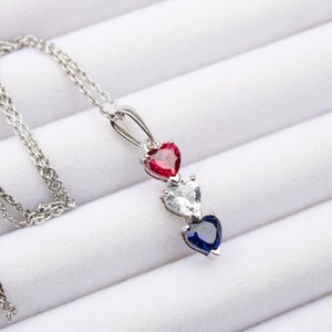 May include: A silver necklace with a pendant featuring three heart-shaped gemstones. The top heart is red, the middle heart is white, and the bottom heart is blue.