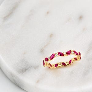 May include: A gold ring with a repeating pattern of marquise-cut ruby gemstones and small, round diamonds. The ring is displayed on a white marble surface, highlighting the red and white stones.