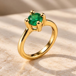 May include: A gold ring featuring a single round-cut vibrant green gemstone held in a four-prong setting. The band is smooth and polished, showcasing a reflective metallic finish that catches the light, highlighting the contrast between the gold and stone.