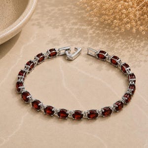 May include: A silver-toned link bracelet featuring a series of oval-shaped, deep red garnet gemstones set in individual metal frames. The bracelet includes a secure metal clasp mechanism and is arranged in a circular shape on a flat surface.